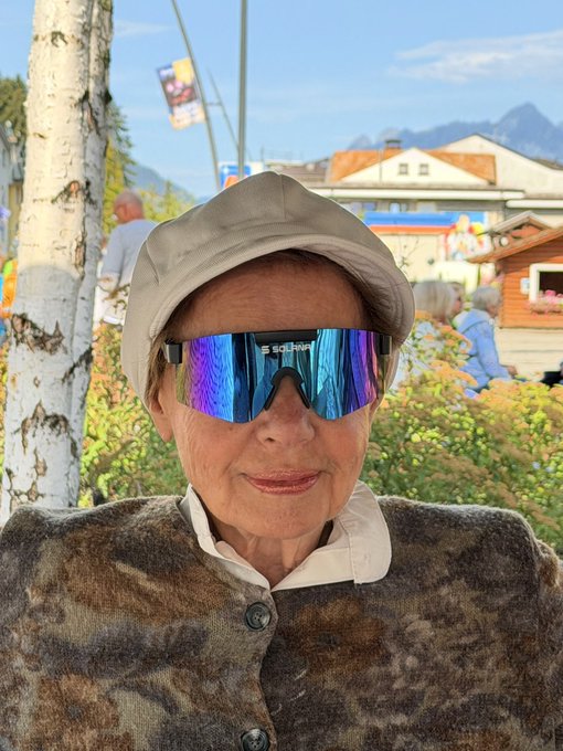 A close-up photo of an elderly person wearing a beige hat and large iridescent blue/purple sunglasses, set against a background of trees, buildings, and a mountain.