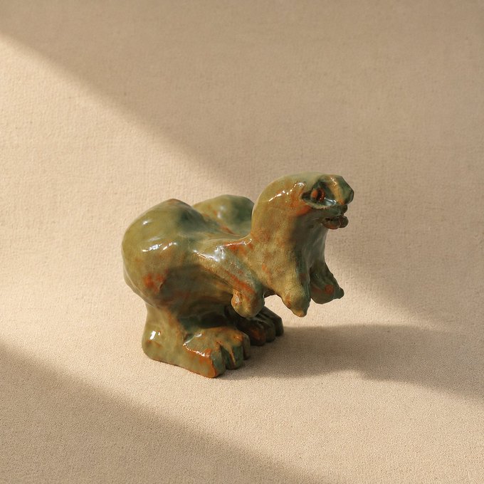 A glazed ceramic figurine of a creature resembling a dinosaur or monster, finished in green and orange tones, sitting on a beige surface with dramatic shadow lighting.