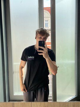 A mirror selfie of a young man wearing a black t-shirt, standing in front of a window with a building visible outside.