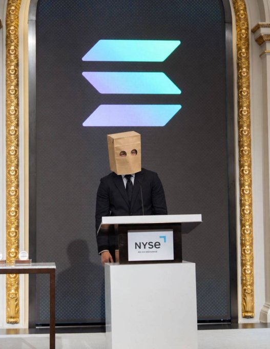 A man in a suit wearing a paper bag over his head stands at a podium marked "NYSE" with the Solana logo displayed on the screen behind him. The image humorously illustrates the post's criticism of Solana's promotional choices, suggesting that backing $ZCAT over $STONK reflects poor judgment—as if even Solana itself is embarrassed to show its face.