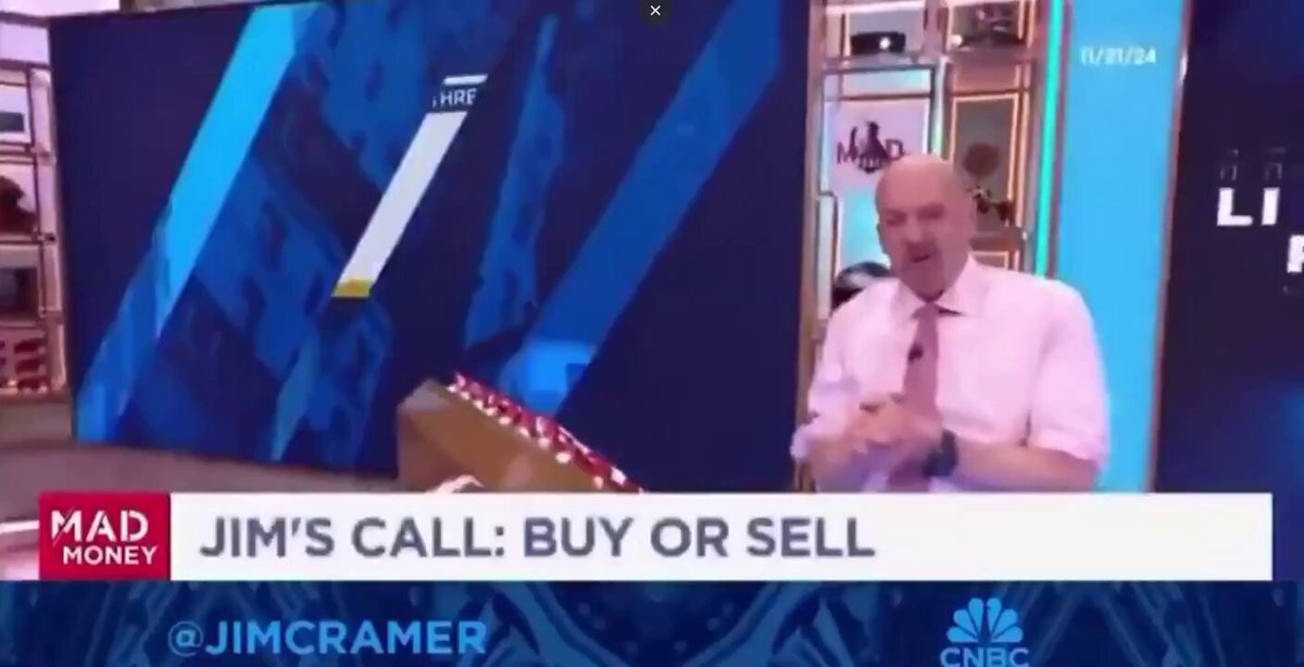 The image features a man sitting at a desk with his hands on his knees while talking into a microphone. The background of the image shows a blue wall, which has an advertisement for Bitcoin written on it. The text "Jim Cramer" is also visible in the image.