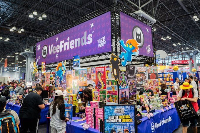 A bustling convention center booth for VeeFriends, featuring large purple signage with cartoon characters and displays of merchandise being browsed by attendees.