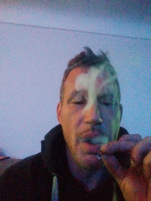 A selfie of a man exhaling smoke against a dusky blue sky, with green and pink hues of light illuminating his face.