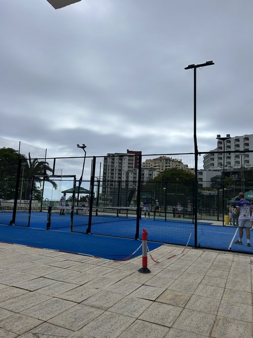 The image shows a tennis court with blue lines, surrounded by tall buildings. There are people playing tennis on the court, while others watch from behind the fence. The sky above appears cloudy, suggesting an overcast day. In the foreground of the photo, there is a red and white cone placed near the net, possibly indicating that someone has just finished their game or practice session.