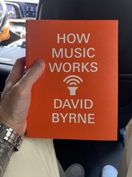 A hand holding a red book titled 'HOW MUSIC WORKS' by David Byrne, photographed inside a car.