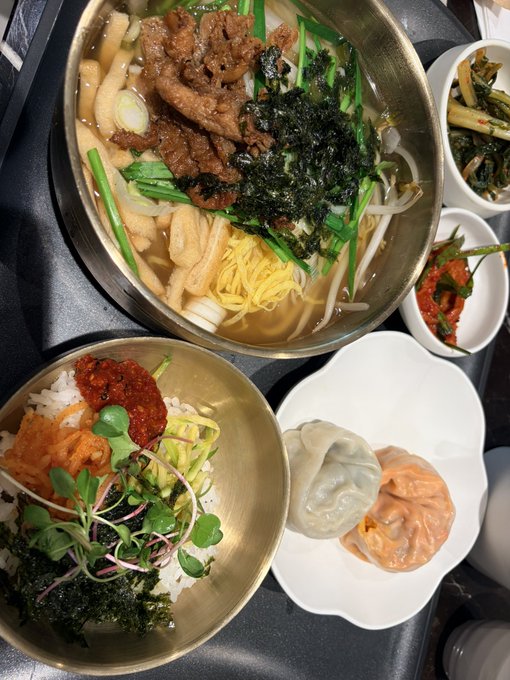 An overhead view of a Korean meal featuring a bowl of soup with noodles, beef, and seaweed, accompanied by rice with gochujang, dumplings, and various side dishes in brass and white bowls.