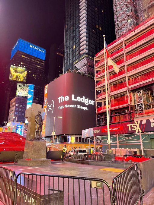 The image shows a city street at night with tall buildings, including one that has a large screen displaying "The Ledger That Never Sleeps". The street appears empty, and there are no people visible in the scene. A fence can be seen on the right side of the photo, possibly separating different areas or enclosing an outdoor event space.