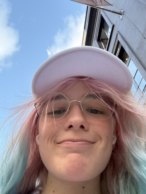A close-up selfie of a young woman with pink hair, round glasses, and a white visor smiling outdoors.