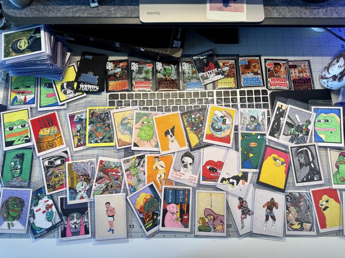 A desk covered in numerous colorful trading cards, stickers, and small prints featuring various cartoon characters, animals, and pop culture images, scattered in front of a keyboard and monitor.