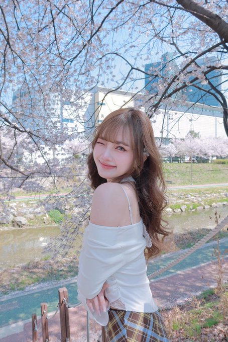 A young woman with long brown hair poses smiling under cherry blossom trees near a stream, wearing a white off-shoulder top and plaid skirt.