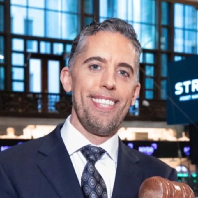 A man in a dark suit and patterned tie smiles while holding a football on what appears to be a sports news set.