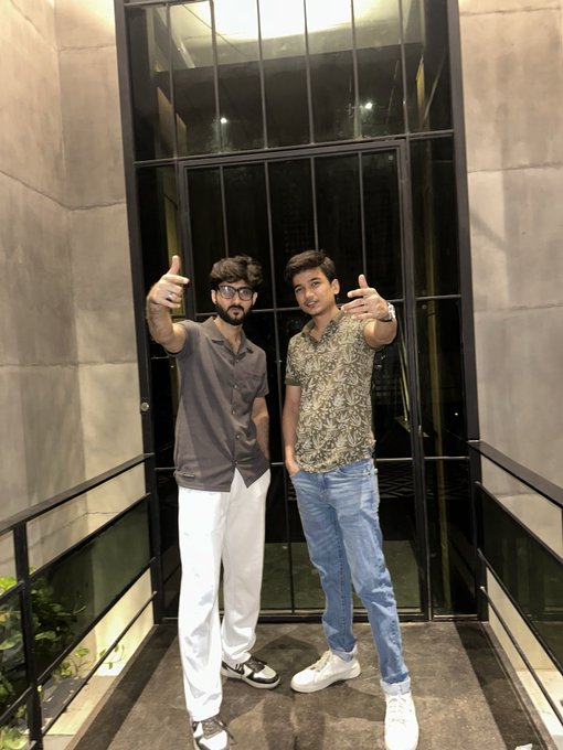 A photograph of two young men posing together on a balcony at night, both pointing their fingers toward the camera in a friendly gesture.