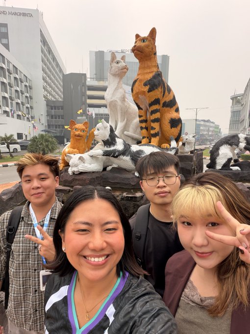 A selfie of four smiling young people posing in front of cat statues on Cat Street in Kuching, Malaysia, with two of them making peace signs.