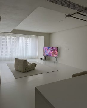 A minimalist modern living room with white walls and a gray floor, featuring a colorful floral painting displayed on a TV stand and a beige bean bag chair.