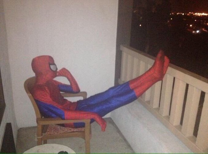 Spider-Man relaxing in a chair on a balcony at night, legs propped up on the railing while patiently waiting to place a bid.