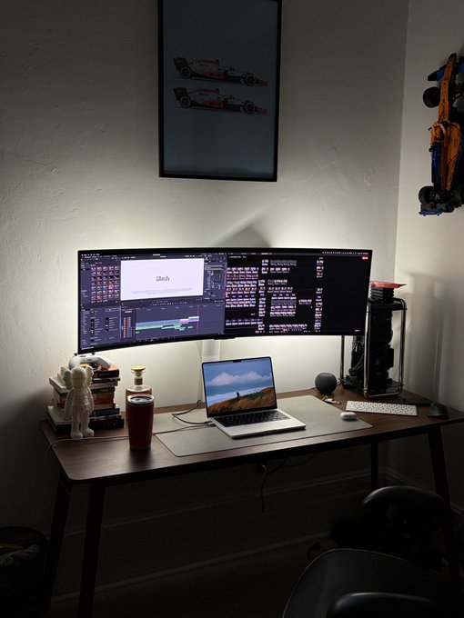 A dimly lit home office setup featuring a triple-monitor workstation where the center screen displays the text "iloh" alongside editing software, with a laptop showing a mountain landscape below—reflecting the creator's intensive 22-hour video editing session. A framed racing poster, astronaut figurine, and PC tower complete the dedicated workspace where the extensive AI-powered edit was completed using Trymelius.