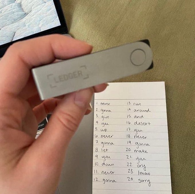 A hand holds a silver Ledger hardware wallet above a notepad listing 24 recovery seed words, illustrating the typical setup process of recording backup phrases after unboxing a new Ledger device.