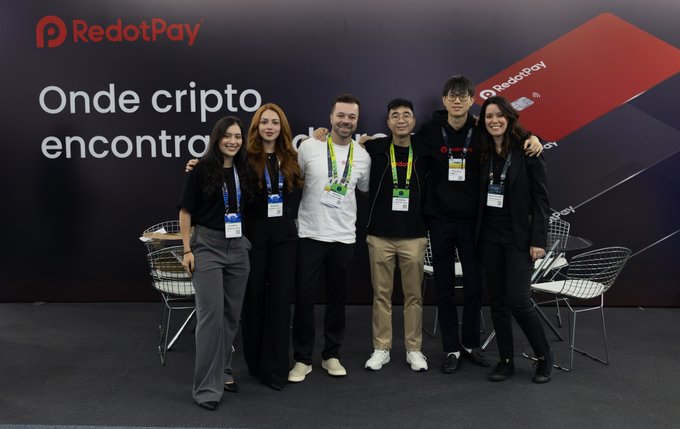 A group of six people wearing RedotPay lanyards pose together in front of a RedotPay-branded backdrop displaying the text 'Onde cripto encontra' (Portuguese for 'Where crypto meets').