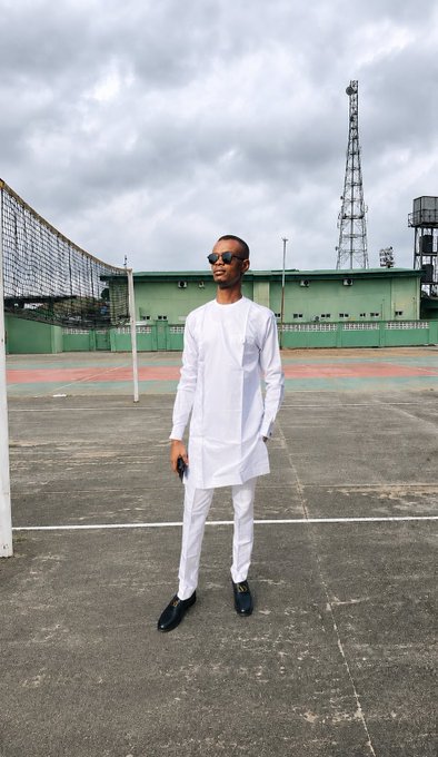 A young man wearing an all-white traditional outfit and sunglasses, standing on an outdoor concrete court with a volleyball net and an industrial facility in the background.