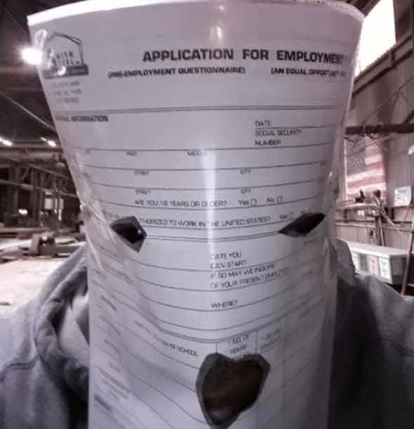 A humorous image of a person wearing a 'APPLICATION FOR EMPLOYMENT' (Pre-Employment Questionnaire) form as a hood, with cutouts for eyes and mouth in a warehouse setting.