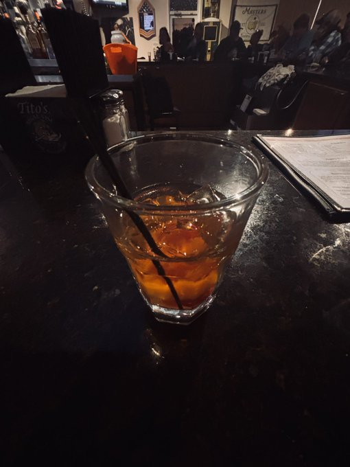 A glass of whiskey on the rocks with a black straw sits on a dark bar counter, with a Tito's Vodka display, a menu, and other patrons visible in the dimly lit background.