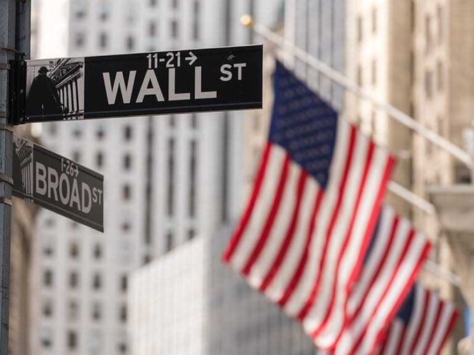 A close-up of Wall Street and Broad Street signs in New York, with American flags waving in the background against tall buildings.