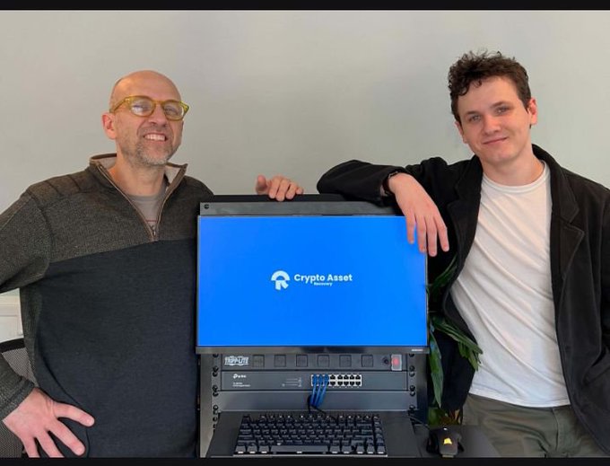 Two men stand beside a Crypto Asset-branded computer or crypto-related machine.