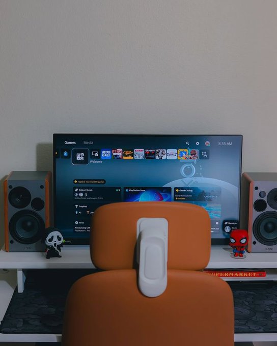 A gaming setup features an orange chair facing a monitor displaying a gaming menu, flanked by Edifier speakers and small Ghostface and Spider-Man figurines, with a book titled 'SUPERMARKET' underneath.