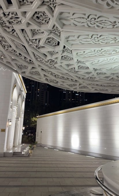 An ornate white coffered ceiling covers a stone walkway leading toward a modern building at night.