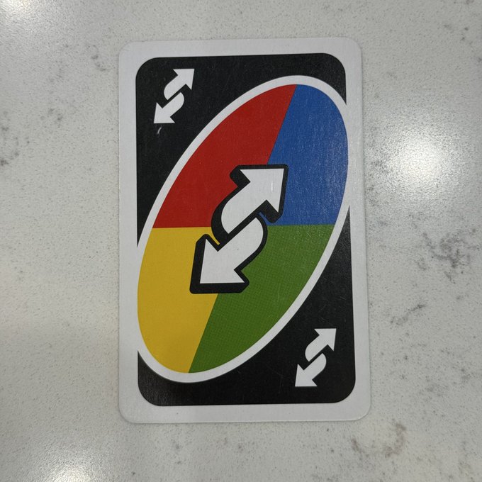 A black playing card featuring a colorful four-quadrant oval design in red, blue, yellow, and green, with white directional arrows including a skip symbol in the center.