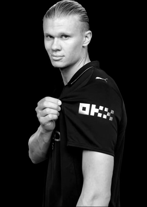 A black-and-white photo of soccer player Erling Haaland pulling at his black Puma jersey to reveal the "OKX" sponsor logo, illustrating his partnership with the cryptocurrency exchange. The image supports the post's message contrasting Haaland's collaboration with OKX against a fictional claim about Mbappé.