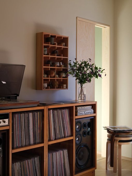 A cozy home interior featuring wooden shelving filled with vinyl records, a small wall-mounted curio shelf with ceramics, a turntable, a speaker, and a vase of purple flowers.