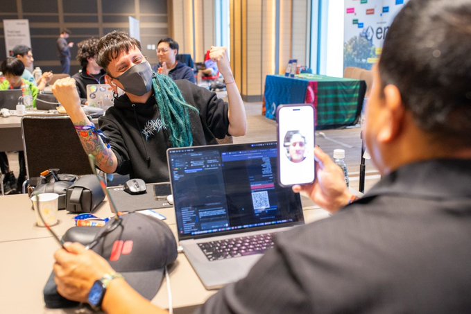 Two people at what appears to be a tech or gaming event, with one person wearing a black hoodie and face mask excitedly raising their fists while another person holds up a phone showing an avatar/face image, with a laptop displaying code or a dashboard in the foreground.