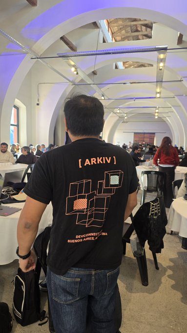 The image features a man wearing a black t-shirt with orange text that says "ARKIV" on it. The man appears to be looking down, possibly observing something or someone behind him. In the background, there are several other people present in the room, suggesting that this gathering might be taking place at an event or conference. There is also a backpack visible near one of the individuals in the scene.
