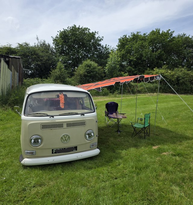 A vintage beige Volkswagen camper van is parked on a grassy field with an orange-striped awning extended, a table and chairs set up nearby, and what appears to be a lei hanging in the windshield.