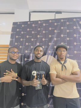Three men pose together in front of a black backdrop covered with 'ASTER' logos, with the central figure holding an ASTER-branded award trophy while wearing a matching ASTER t-shirt.