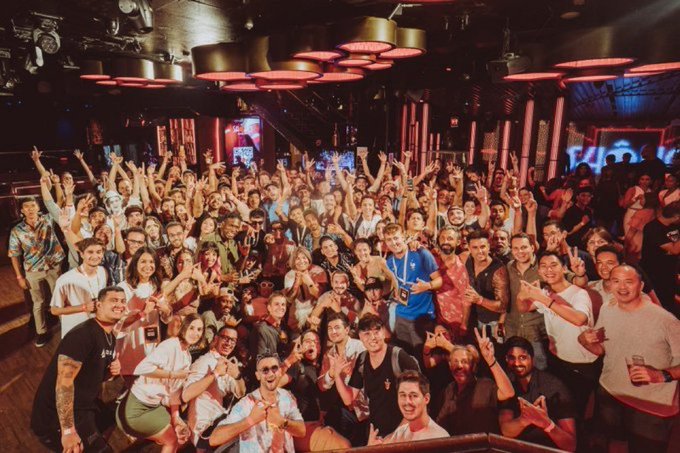 A large group of people gathered in a dimly lit club or venue with red lighting, raising their hands and smiling for a group photo.