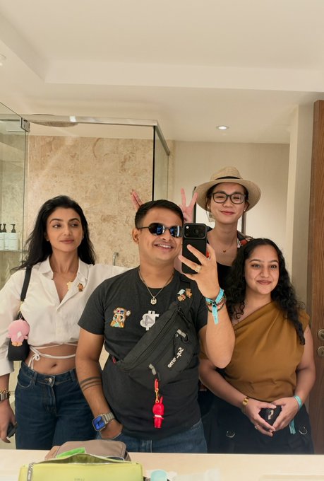 A group of four people taking a mirror selfie in what appears to be a bathroom, including a man with sunglasses and a cross-body bag, and a woman in a straw hat.