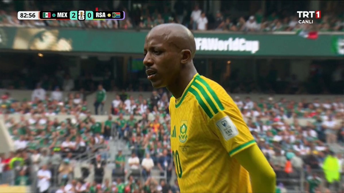 The image shows a soccer player wearing a yellow jersey walking across the field, with a crowd of people watching him from behind. The player appears to be focused on his game, possibly preparing for an upcoming play or discussing strategies with teammates. In the background, there is a scoreboard displaying the current score and time remaining in the match.