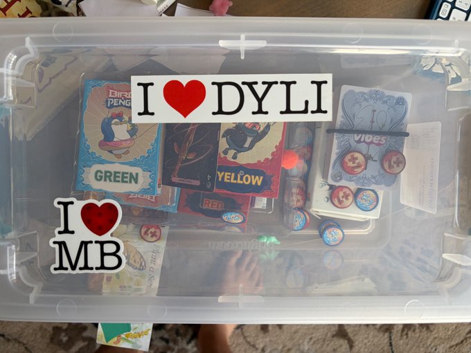 A photograph of a clear plastic storage container holding collectible items, with text stickers reading 'I ❤ DYLI' and 'I ❤ MB' placed on top of card decks and chips.