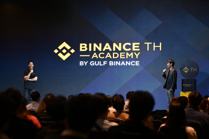 The image shows a large crowd of people gathered for a presentation by two men standing behind a podium with a blue backdrop featuring the words "Binance TH Pizza Day" written above them. The audience appears engaged, as they are watching the speakers intently. The event seems to be focused on discussing the future direction and potential growth opportunities in trading card games (TCG) and real-world assets.