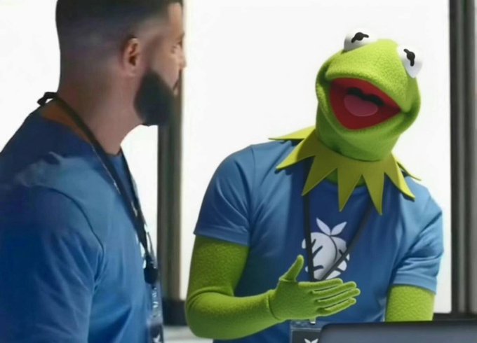 A man in a blue shirt stands next to Kermit the Frog wearing a blue t-shirt with an apple logo and a yellow spiked collar, appearing to be in an office setting.