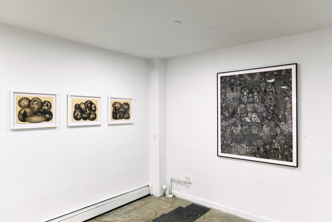 A minimalist white-walled art gallery showing three small framed pieces of dark, clustered forms on the left and one large framed artwork with intricate, surreal dark imagery on the right.
