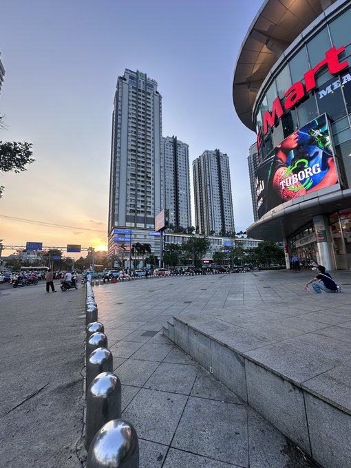 A bustling urban plaza at dusk features a "VinMart" store with a prominent "Tuborg" advertisement, surrounded by tall high-rise residential towers and a row of shiny metal bollards, with people scattered across the open square. The image's depiction of a vibrant, active cityscape complements the post's excitement about the crypto market surging green across multiple coins like ZEC, HYPE, TRUMP, SOL, and ETH.