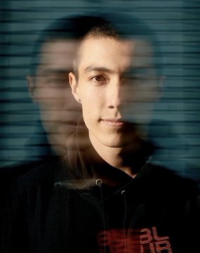A young man with short hair poses for a portrait shot through window blinds, wearing a black hoodie with partial red and white text reading "BLAST" and "PAC." The image accompanies a post detailing the biography of Blast and Pacman, a self-taught programmer and Thiel Fellowship recipient who founded the decentralized domain platform Namebase.