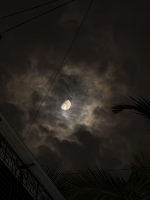 A nighttime photograph of a crescent moon shining through scattered clouds, with power lines, a building edge, and palm fronds silhouetted in the foreground.