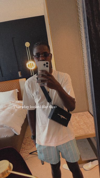 A man in a white t-shirt and striped shorts takes a mirror selfie in a hotel room, wearing sunglasses and a crossbody bag, with text 'Thursday looked like this' overlaid.