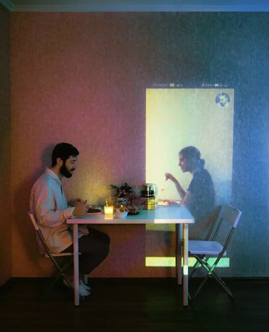 An artistic photo of a man sitting at a table across from a projected video call showing a woman, with drinks and a candle between them.