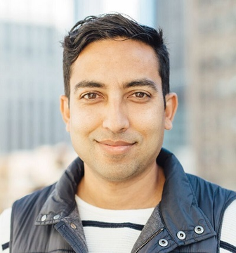 A smiling man in a dark quilted vest over a striped shirt poses for a close-up portrait against a blurred urban backdrop. The image likely accompanies the post featuring Evernode CEO Billa, who commented on AI agents settling RLUSD transactions on the XRP Ledger without human involvement.