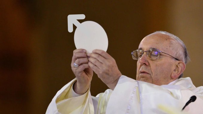 A photograph of Pope Francis in white robes, holding up a white circular communion host with an overlaid male symbol (arrow pointing up-left) in the upper left corner.